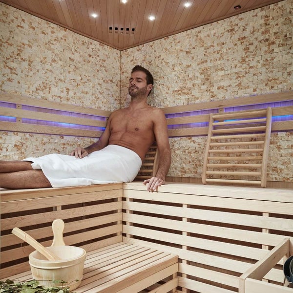Eine Person gießt mit einer Holzkelle Wasser auf die heißen Saunasteine eines elektrischen Saunaofens in einer hellen Holzsauna.