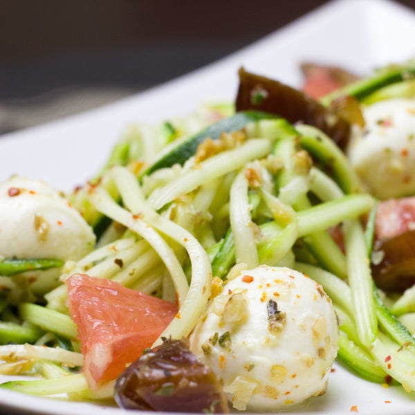 Salat mit Zucchini-Nudeln, Mozzarella-Bällchen, Tomatenstücken und Zitrusfrüchten, garniert mit Kräutern und Nüssen auf einem weißen Teller.