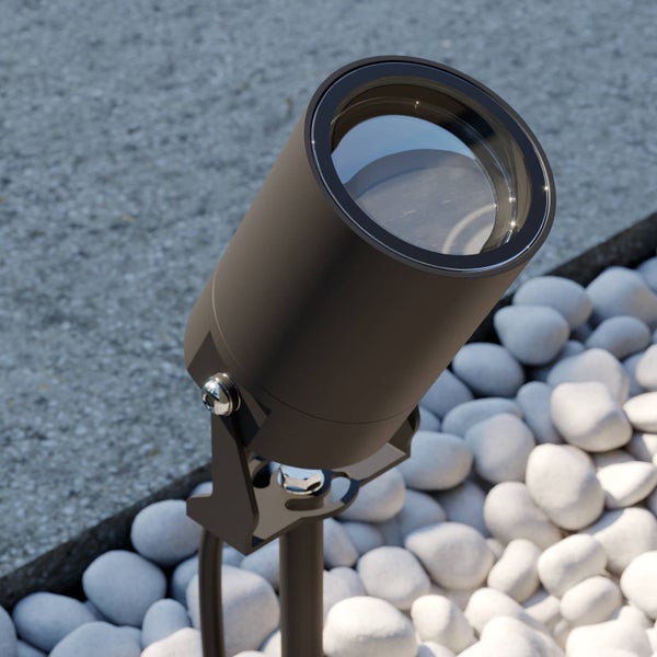 Moderner verstellbarer Außenstrahler aus dunkelbraunem Metall, montiert in einem Gartenbeet mit weißen Kieselsteinen und Betonkante.