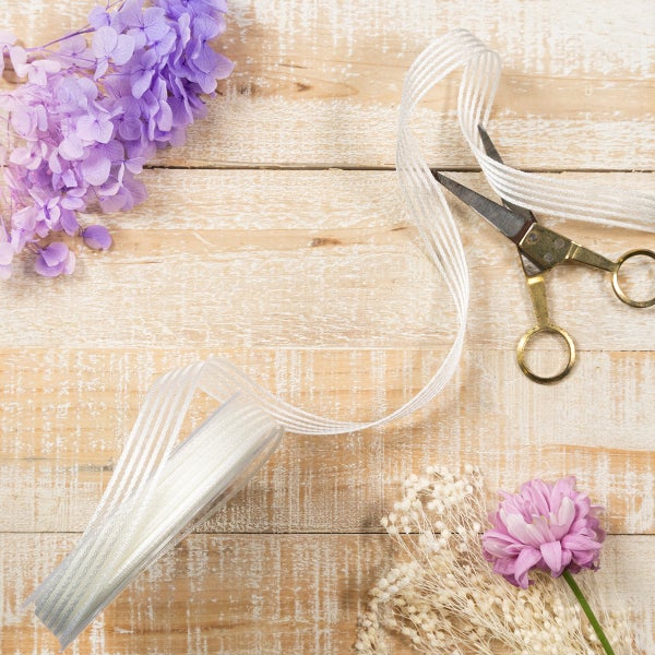 Floristen-Set auf Holzgrund mit weißem Dekoband, goldfarbener Schere und lila Blumen für kreatives Basteln und Dekorieren.