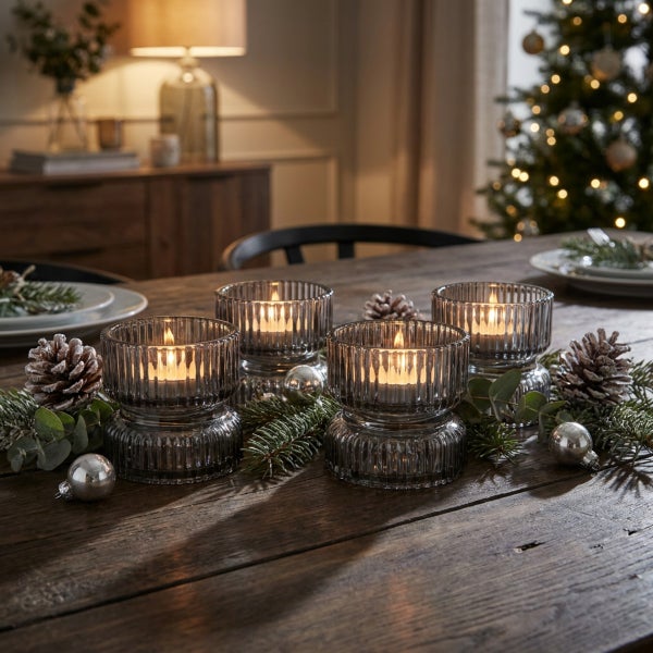 Vier gerippte Windlichter aus Glas mit brennenden Teelichtern auf einem Holztisch dekoriert mit Tannenzweigen, Zapfen und Weihnachtsschmuck.