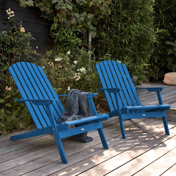 Zwei blaue Adirondack-Gartenstühle aus Holz auf einer Holzterrasse, umgeben von grünen Pflanzen und weißen Rosen.