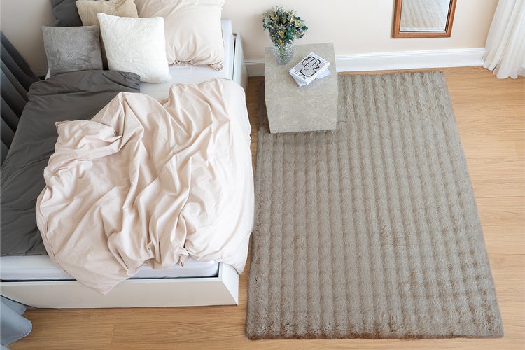 Ein rechteckiger Teppich mit strukturierter Oberfläche liegt neben einem Bett in einem modern eingerichteten Schlafzimmer mit Holzfußboden.