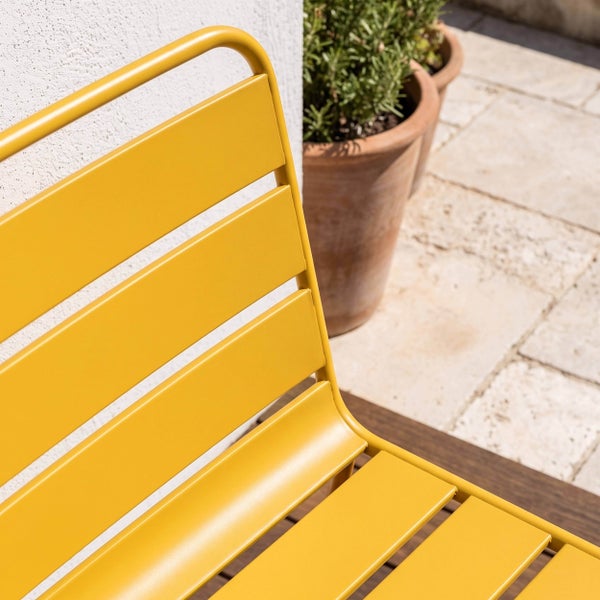 Nahaufnahme eines Gartenstuhls aus Metall mit Lamellen auf einer Terrasse mit Pflanzkübeln.