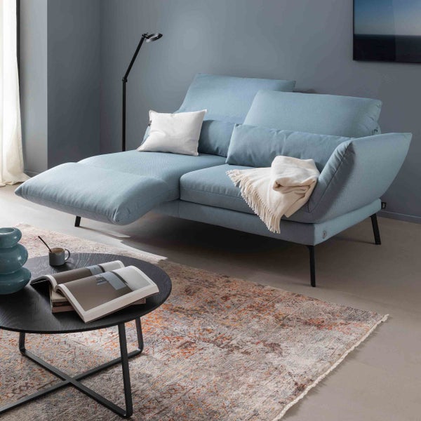 Modernes hellblaues Sofa mit verstellbaren Kopfstützen und Fußstütze in einem Wohnzimmer mit rundem Couchtisch und gemustertem Teppich.