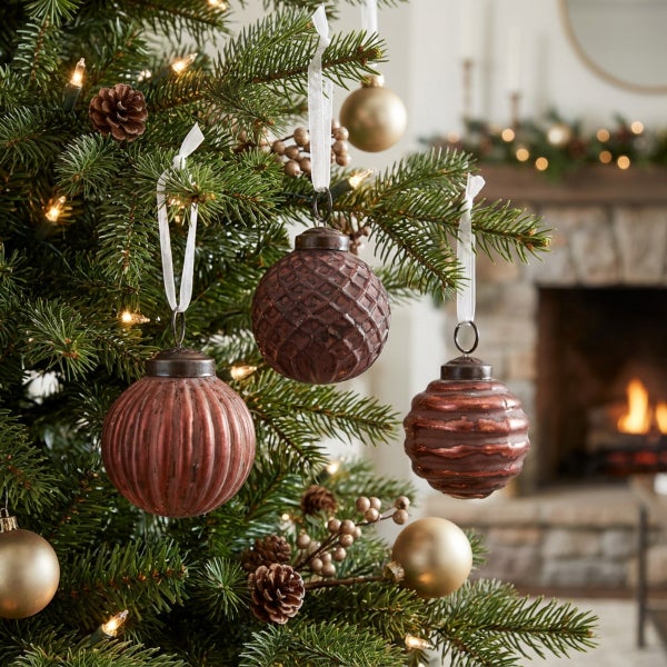Set mit strukturierten Weihnachtsbaumkugeln aus Glas an einem Tannenbaum vor einem Kamin in rustikaler Wohnumgebung.