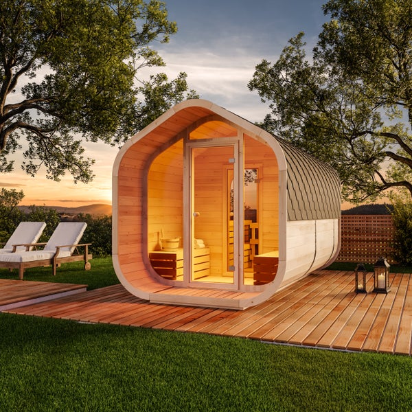 Ovale Außensauna aus Holz mit Glasfront auf einer Holzterrasse im Garten bei Sonnenuntergang.