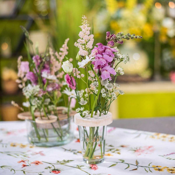 Glasvasen mit Holzeinsätzen für Blumenarrangements auf einer Tischdecke mit Blumenmuster, dekoriert mit Löwenmäulchen, Astilbe und Sommerblumen.
