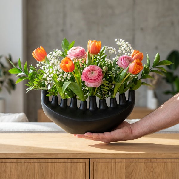 Hand hält eine schwarze Blumenvase mit einer Anordnung aus Tulpen, Ranunkeln und Schleierkraut auf einem Holztisch.