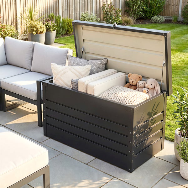 Geöffnete Garten-Auflagenbox aus Metall in Anthrazit, gefüllt mit Kissen und Stofftieren auf einer Terrasse neben einem Loungesofa.