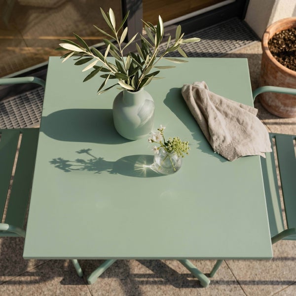 Quadratischer Gartentisch aus Metall in Hellgrün mit passenden Stühlen, dekoriert mit Olivenzweigen und einer Serviette auf einer Terrasse.