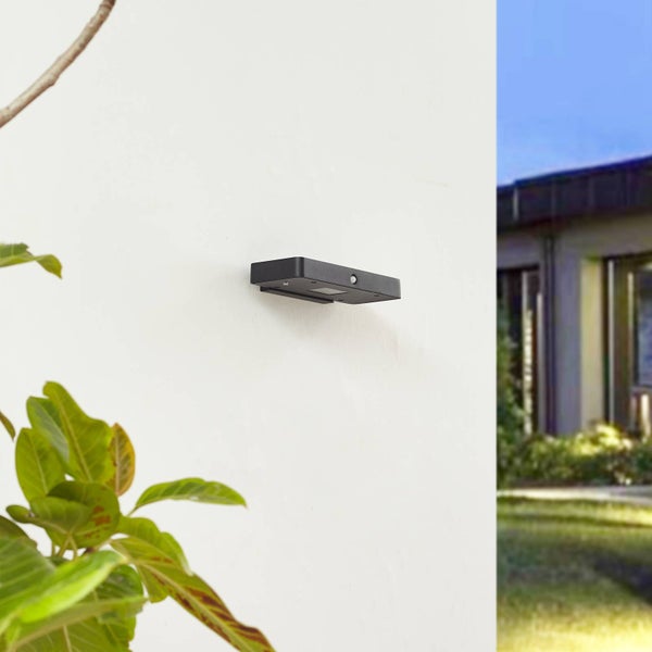Moderne rechteckige Außenwandleuchte mit Bewegungsmelder, montiert an einer weißen Fassade in einer hellen Gartenumgebung.