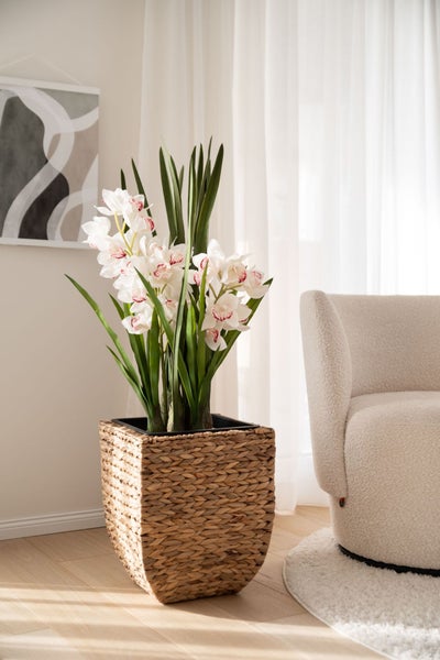 Künstliche weiße Orchidee in einem geflochtenen quadratischen Korb, platziert in einem hellen, modernen Raum neben einem hellen Sessel.
