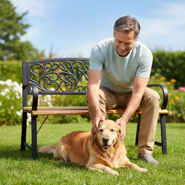 Ein Mann sitzt auf einer Gartenbank aus Metall und Holz und streichelt einen Golden Retriever auf einer Rasenfläche im Garten.