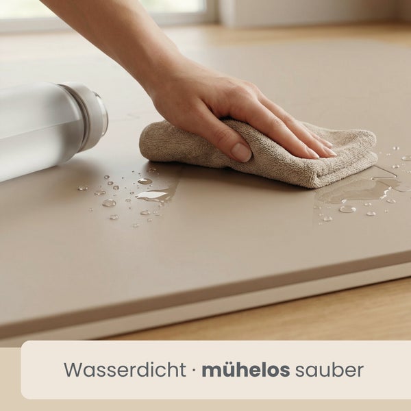 Eine Hand wischt mit einem Tuch Wasser von einer wasserdichten Matte auf einem Holzboden ab.