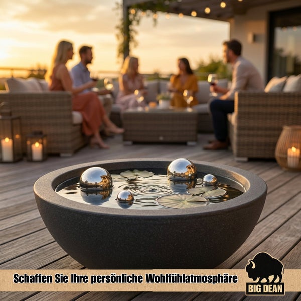 Runde Wasserschale mit dekorativen Edelstahlkugeln und Seerosenblättern auf einer Terrasse, im Hintergrund Menschen in geselliger Runde.