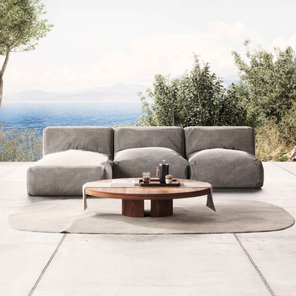 Modernes graues Outdoor-Sofa mit rundem Holztisch und ovalem Outdoorteppich auf einer hellen Steinterrasse mit Meerblick.