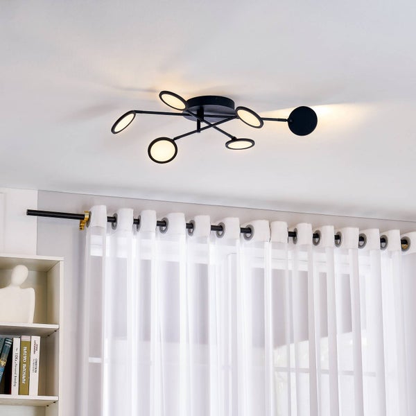 Moderne schwarze Deckenleuchte aus Metall mit runden Leuchtelementen in einem hellen, minimalistischen Raum mit Vorhang und Bücherregal.