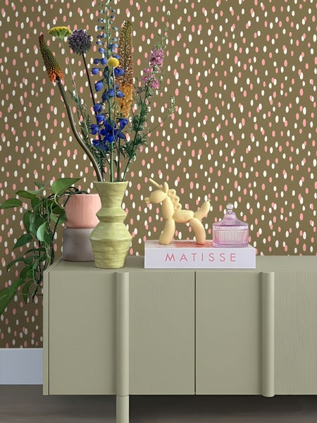 Dekoratives Sideboard mit Blumen, Pflanzen, Buch und Dekorationsfiguren vor einer gepunkteten Tapete