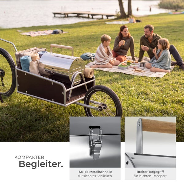 Tragbarer Edelstahlgrill im Transportwagen bei einem Familienpicknick am See, mit Detailaufnahmen von Metallschnalle und Holzgriff.