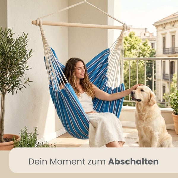 Eine Frau sitzt entspannt in einem blau gestreiften Hängesessel aus Stoff auf einem hellen Balkon neben einem Hund und Pflanzen.