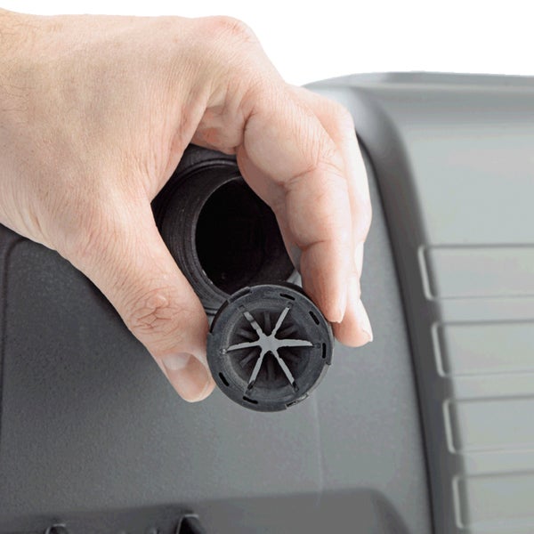 Hand hält runde schwarze Kunststoffdichtung mit sternförmiger Öffnung über den Einlass eines grauen Kunststoffbehälters.