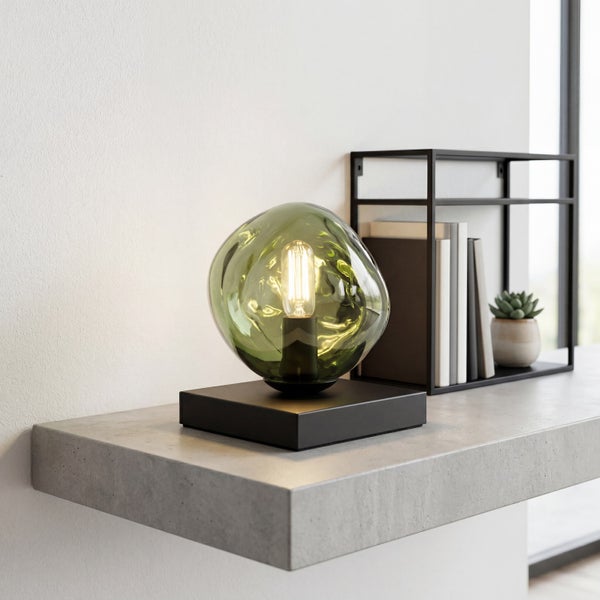 Tischleuchte mit grünem Glasschirm in organischer Form und schwarzem Sockel auf einem Betonregal.