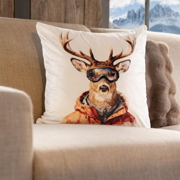 Dekokissen mit Hirschmotiv, der eine Skibrille und Skijacke trägt, auf einem Sofa in einem Raum mit Blick auf schneebedeckte Berge platziert.