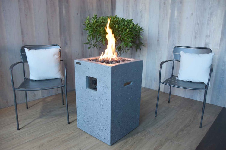 Quadratische Gasfeuerstelle in Säulenform aus Verbundwerkstoff mit zwei Metallstühlen, Kissen und einer Pflanze vor einer Holzverkleidung.