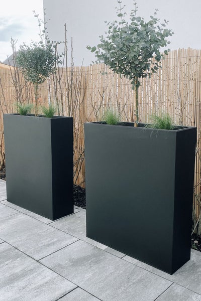 Zwei hohe rechteckige anthrazitfarbene Pflanzgefäße auf einer Terrasse mit Natursteinplatten vor einer Sichtschutzmatte aus Schilfrohr.