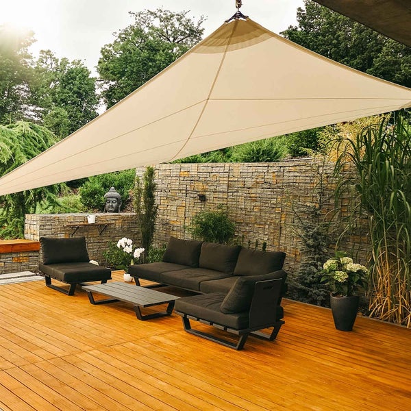 Großes helles Sonnensegel über einer Holzterrasse mit dunklen Lounge-Gartenmöbeln und einer Steinwand im Hintergrund.