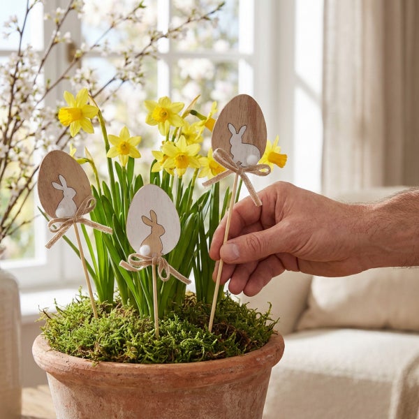 Hand steckt hölzerne Osterstecker mit Hasenmotiv in einen Tontopf mit Narzissen und Moos als frühlingshafte Dekoration.