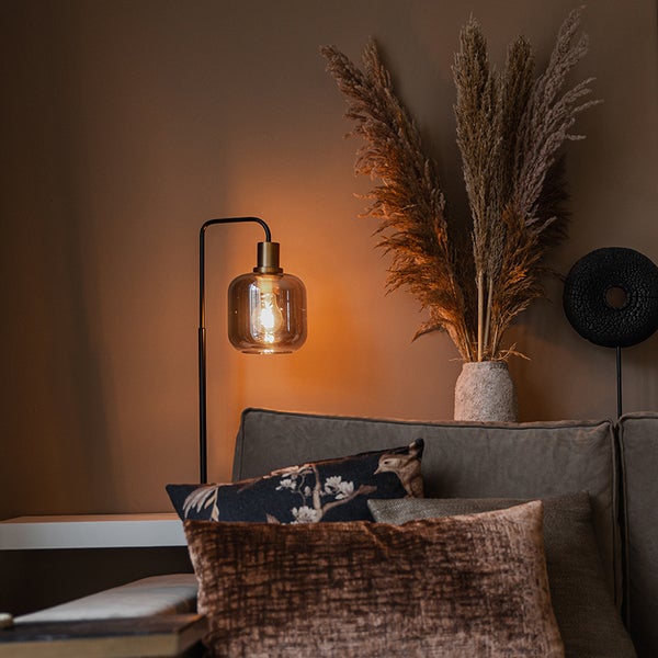 Gemütlich eingerichteter Wohnbereich mit einer Stehleuchte aus Metall und Glas, einem Sofa mit strukturierten Kissen und einer Vase mit Pampasgras.