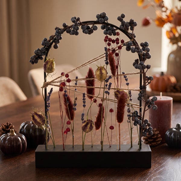Tischdekoration im Herbstdesign mit Trockenblumen und Beeren an einem Metallbogen auf einem schwarzen Sockel auf einem Holztisch.