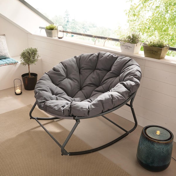 Grauer Papasan-Schaukelsessel mit gestepptem Kissen auf einem hellen Balkon mit Pflanzen, Teppich und Laternen.