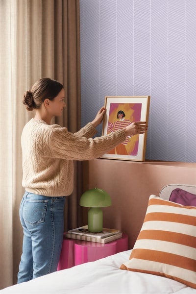 Eine Frau hängt ein gerahmtes Bild in einem hellen Zimmer mit violetter Fischgrät-Tapete, einer grünen Lampe und gestreiften Kissen auf.