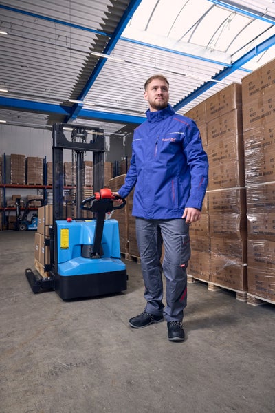 Ein Mann in einem Lager trägt eine blaue Arbeitsjacke, graue Arbeitshose und schwarze Sicherheitsschuhe neben einem blauen elektrischen Hochhubwagen.