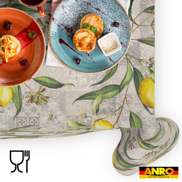 Abwaschbare Wachstuch-Tischdecke mit Zitronen-Motiv und Patchwork-Muster auf gedecktem Tisch, lebensmittelecht, ANRO DEUTSCHE MANUFAKTUR Logo.