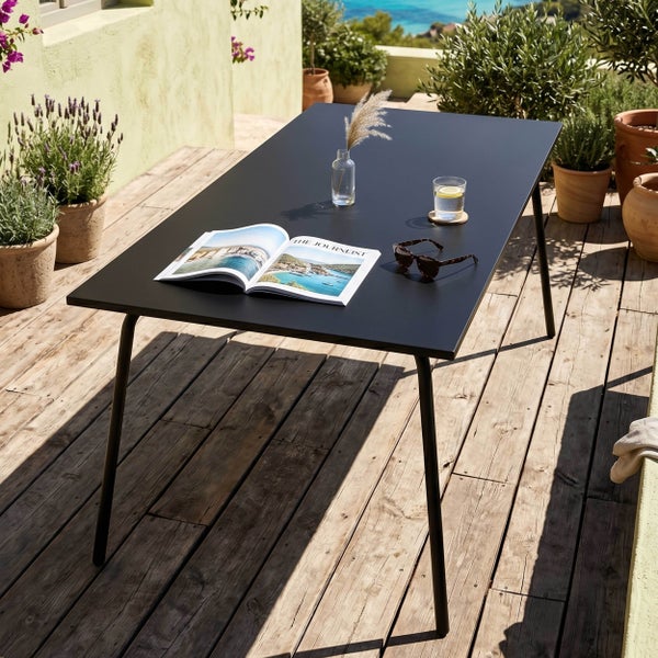 Rechteckiger schwarzer Gartentisch auf einer Holzterrasse, dekoriert mit Magazin und Glas, in heller mediterraner Umgebung mit Pflanzen.