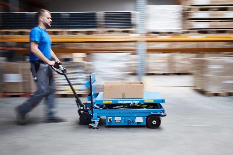 Mann in Arbeitskleidung schiebt einen blauen HanseLifter Scherenhubwagen mit einem Karton durch ein gut organisiertes Lager mit Hochregalen.