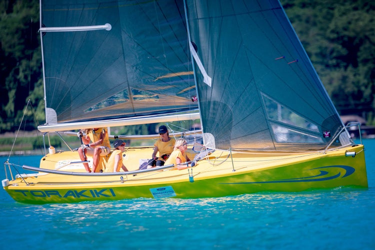 Ein gelbes Segelboot mit Besatzung in gelber Kleidung segelt auf einem See vor einer bewaldeten Kulisse.