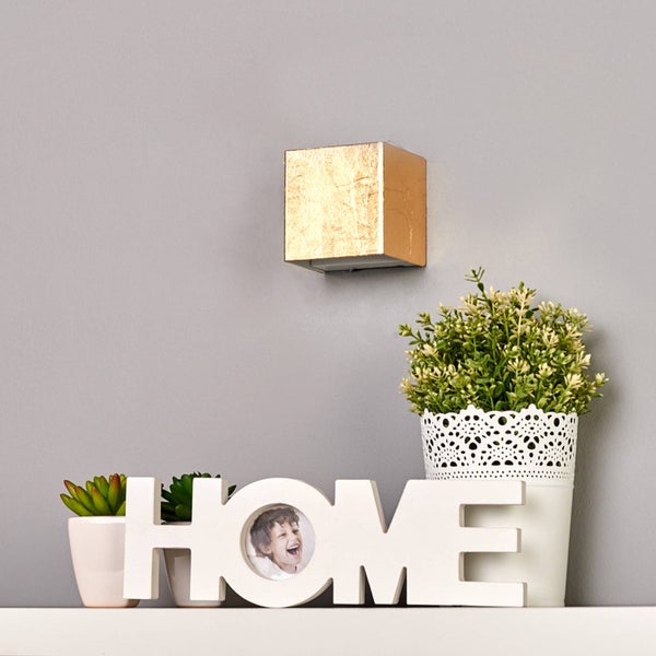 Würfelförmige Wandlampe mit goldener Struktur über einem Regal mit Zimmerpflanzen und einem dekorativen Home-Schriftzug.