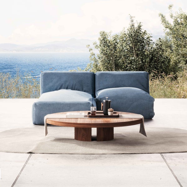 Moderner Außenbereich mit blauem Zweisitzer-Sofa, rundem Couchtisch aus Holz und Teppich auf einer hellen Terrasse mit Meerblick.