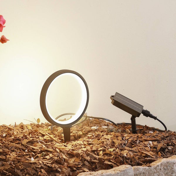 Solarbetriebene LED-Gartenleuchte in Ringform mit separatem Solarmodul auf Erdspießen, platziert in Rindenmulch und leuchtend.