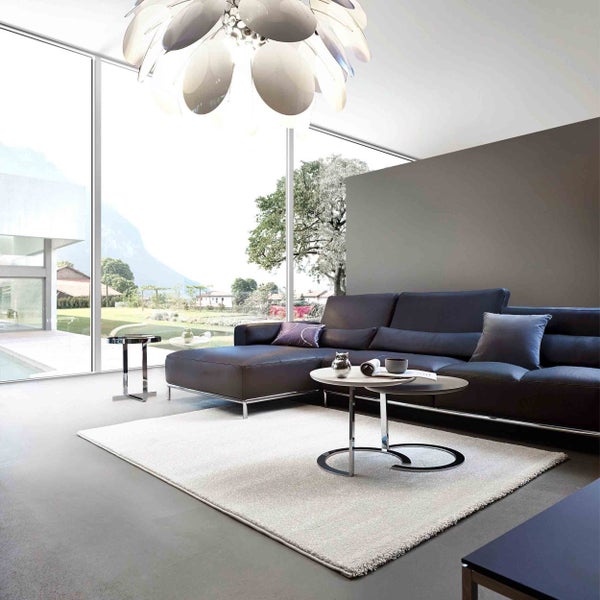 Modernes Wohnzimmer mit dunklem Ecksofa aus Leder, hellem Teppich, runden Couchtischen und großer Fensterfront mit Blick in die Natur.