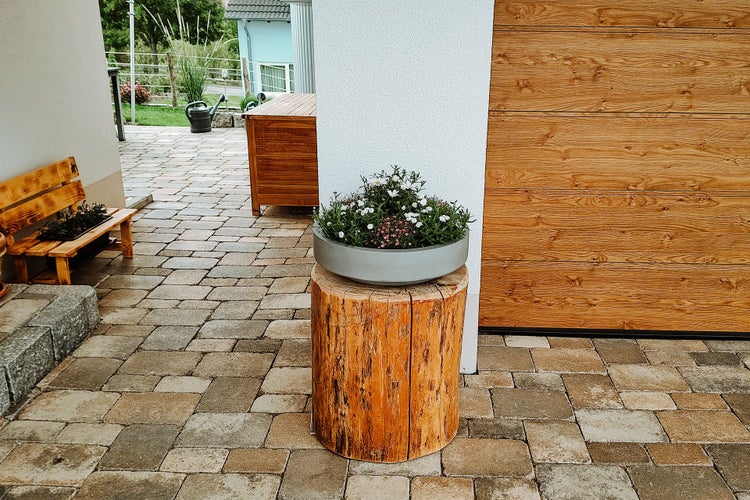 Helle, gepflasterte Terrasse mit einer grauen Pflanzschale auf einem Baumstamm-Podest vor einer Holzwand.