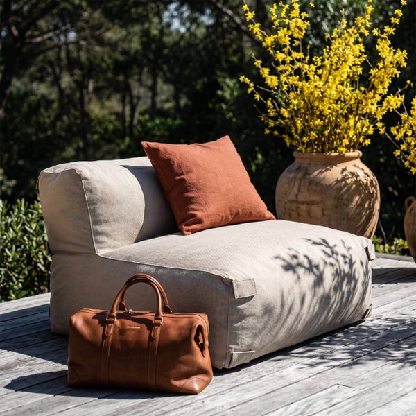 Moderner Outdoor Loungesessel mit terrakottafarbenem Kissen auf einer Holzterrasse neben einer Ledertasche und einem großen Blumentopf.