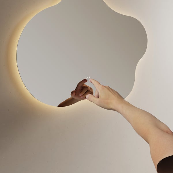 Asymmetrischer LED-Badezimmerspiegel mit Touch-Sensor und indirekter Beleuchtung, eine Hand betätigt den Schalter auf der Spiegelfläche.