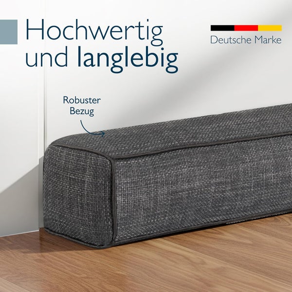 Grauer Zugluftstopper mit robustem Textilbezug von einer deutschen Marke auf einem Holzfußboden an einer Wand.
