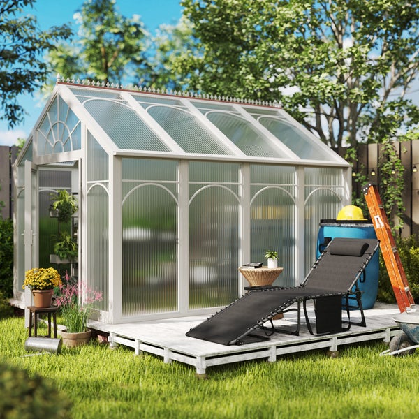 Viktorianisches Gewächshaus mit weißem Rahmen und geriffelten Paneelen auf einem Holzdeck im Garten mit Sonnenliege und Pflanzen.